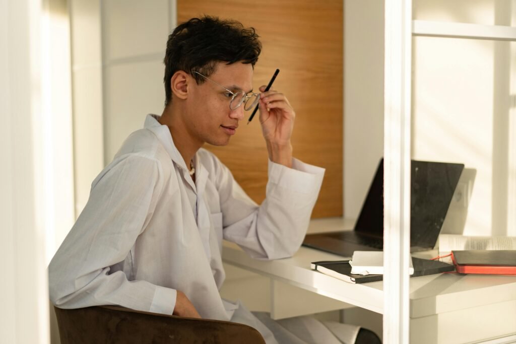Installations A young man wearing a white thobe sits indoors, pondering at a desk with a laptop and notes.