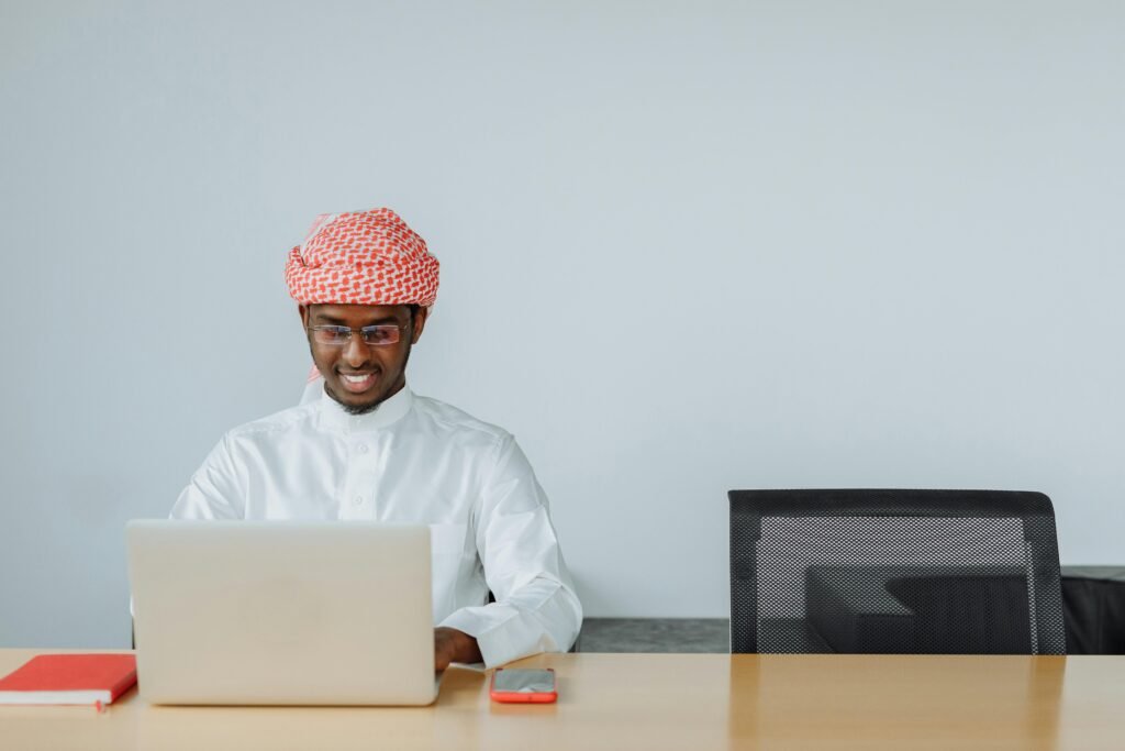 Admissions (process) Middle Eastern man in traditional attire using a laptop at his office desk, smiling while working.